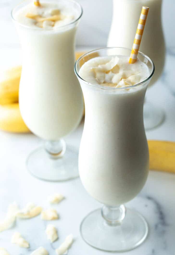 3 große Mixgetränkegläser mit Bananen-Smoothie und einem gelb gestreiften Strohhalm in jedem Glas.