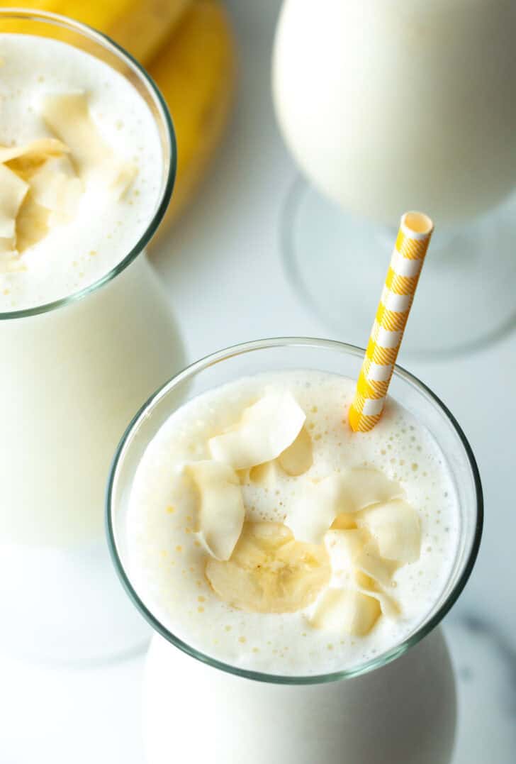 Draufsicht auf ein großes Mixgetränkglas mit Bananen-Smoothie und einem gelb gestreiften Strohhalm in jedem Glas.