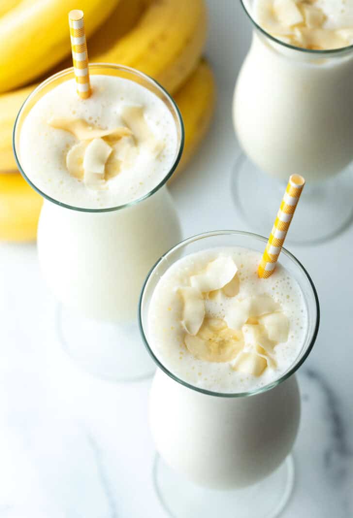 Draufsicht auf ein großes Mixgetränkglas mit Bananen-Smoothie und einem gelb gestreiften Strohhalm in jedem Glas.