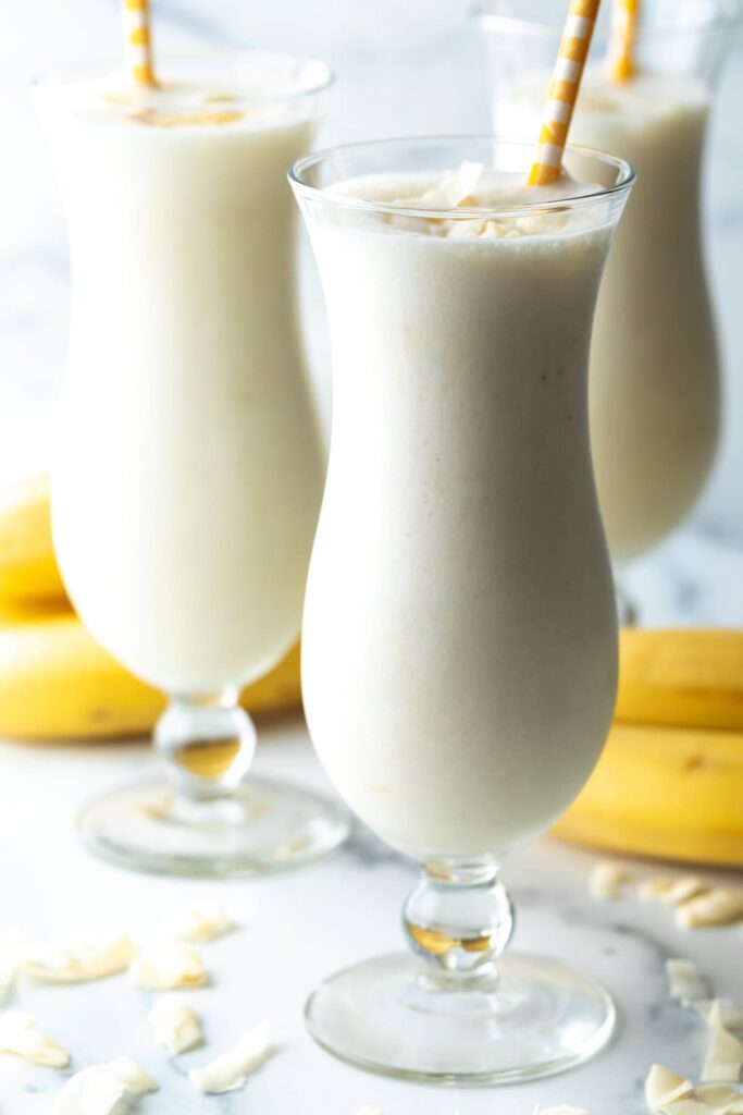 3 large mix drink glasses with banana smoothie and a yellow striped straw in each glass.