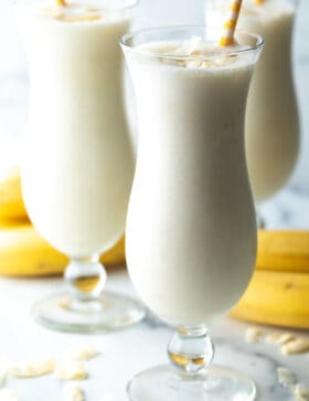 3 große Mixgetränkegläser mit Bananen-Smoothie und einem gelb gestreiften Strohhalm in jedem Glas.