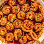 honey garlic shrimp in a white skillet with a wooden spoon holding 1 shrimp. the shrimp are garnished with green onions and white sesame seeds