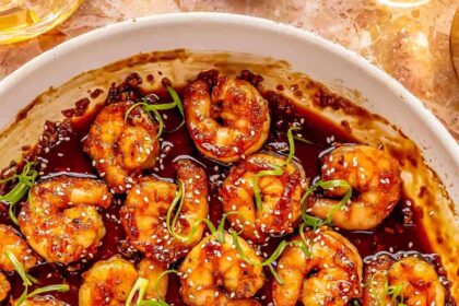 honey garlic shrimp in a white skillet with a wooden spoon holding 1 shrimp. the shrimp are garnished with green onions and white sesame seeds