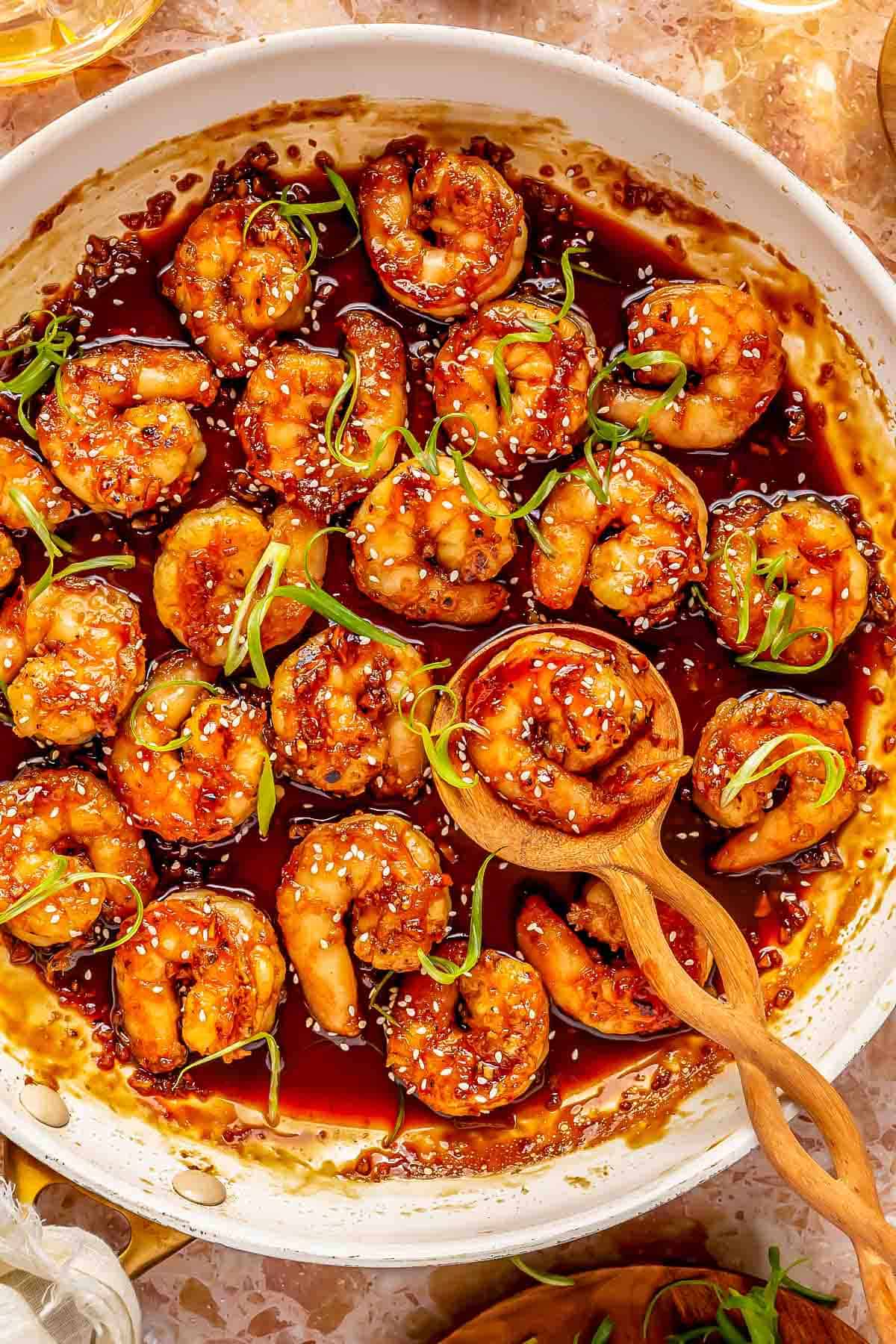 honey garlic shrimp in a white skillet with a wooden spoon holding 1 shrimp. the shrimp are garnished with green onions and white sesame seeds