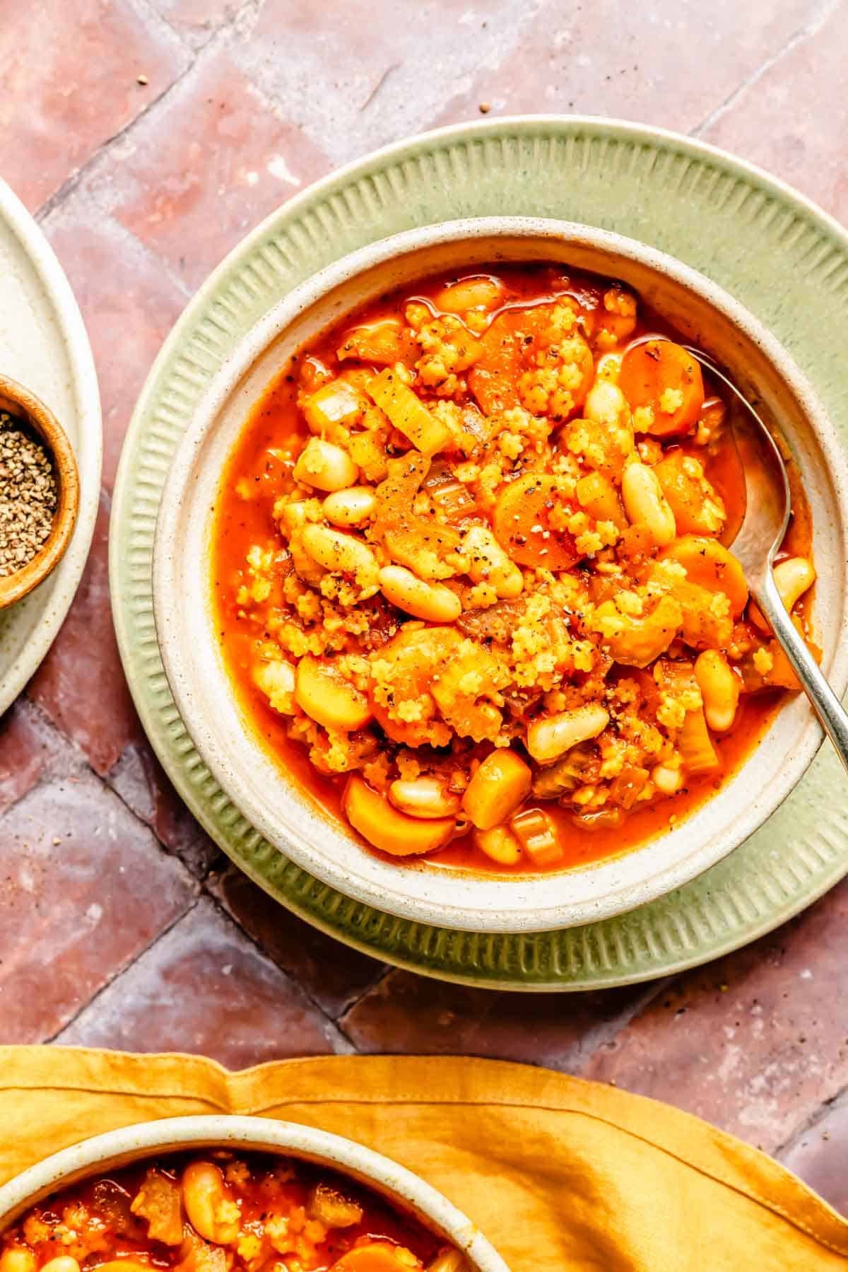 Minestrone-Suppe in einer weißen Schüssel auf einem grünen Keramikteller neben einem gelben Leinenhandtuch